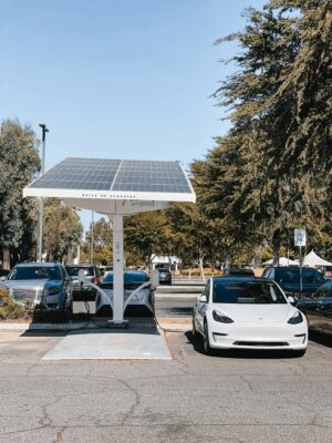 A white electric car charging under a solar panel in an urban setting, showcasing renewable energy use.